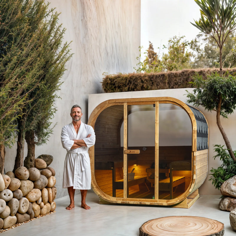Man in a white robe standing next to a wooden barrel sauna in a natural setting with plants and rocks.