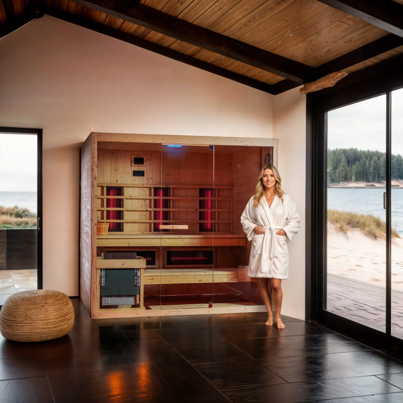 Woman in a white robe standing in front of a Dharani Combi spruce sauna in a room with large windows overlooking a scenic view.