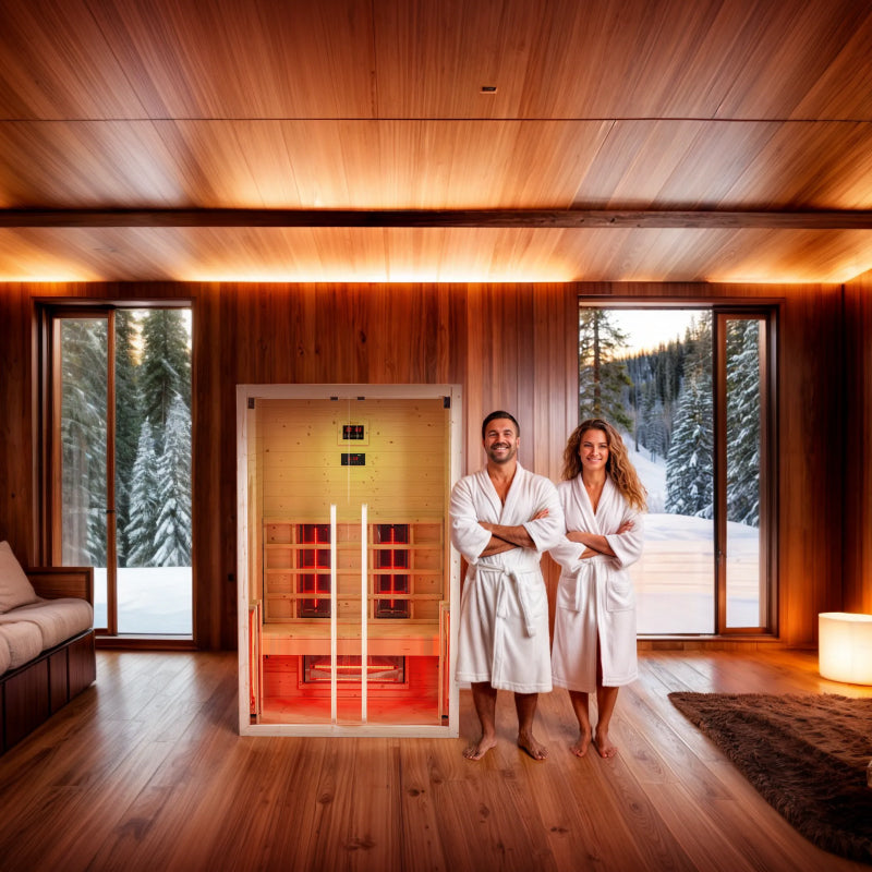 Two people in white robes standing in front of Dharani S2 infrared sauna in a room with wooden interior and large windows showing a snowy landscape.